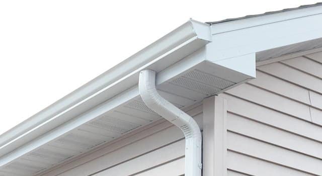 A close-up view of a house's white gutters and downspout system attached to beige vinyl siding under the roof eaves.